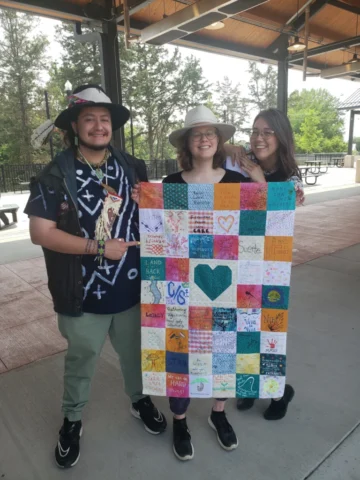 20230616_143943 Three people hold up a colorful quilt with a heart block in the middle that they made as part of Sewing Machine project