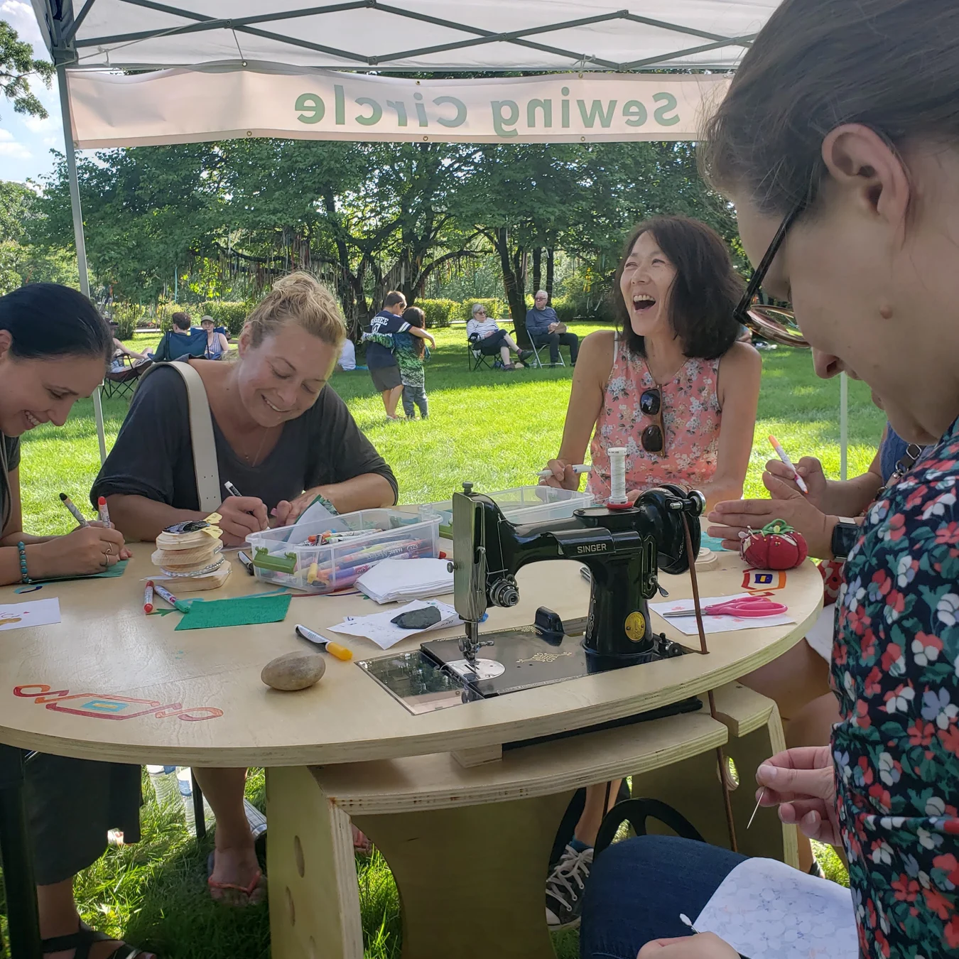 Women gather to draw and sew beneath a tent outdoors.