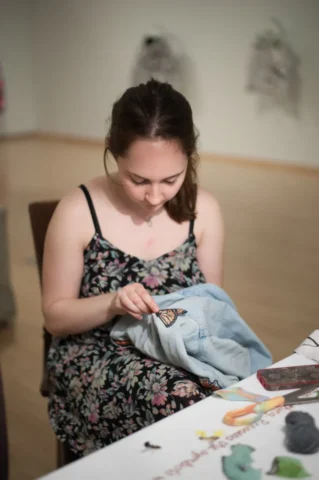 person in a floral dress sews a monarch butterfly patch onto a denim shirt.