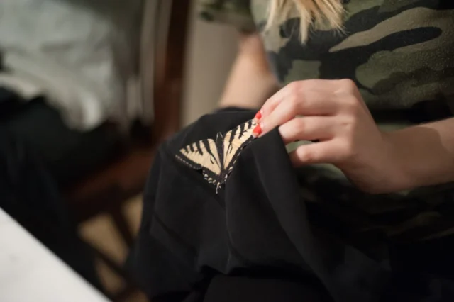 Alterations_Alterations-163 copy a hand with chipped red nail polish positions a swallowtail butterfly patch on a black garment