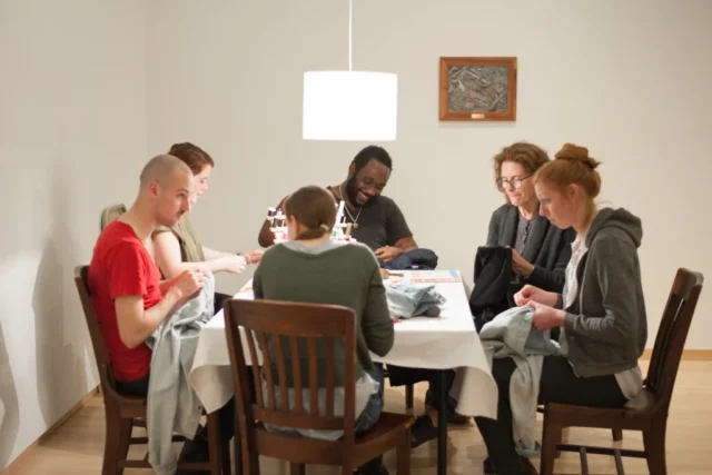 Alterations_Alterations-39 copy a group of people sits around a table, sewing