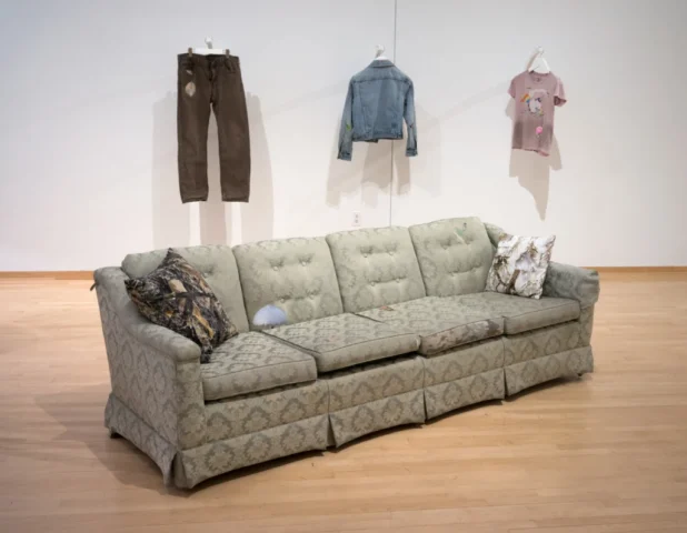 a mended couch sits in a gallery in front of a wall with three garments hanging on the wall.