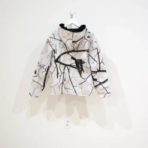 a camouflage, fleece-lined jacket with photos of plastic bags stuck in the dark tree branches against a white background.