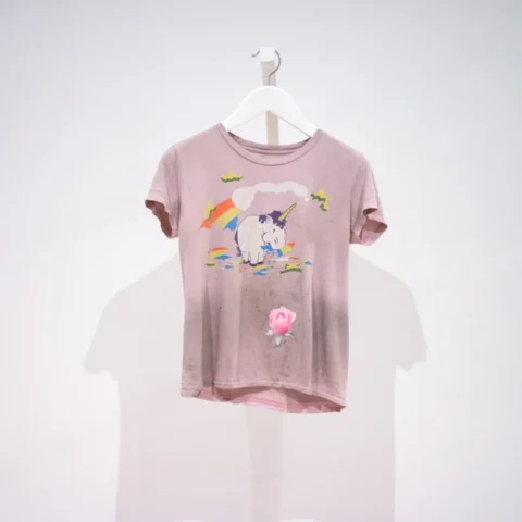 a very dirty pink t-shirt with a graphic of a unicorn eating a rainbow on it. It also has a vibrant light pink rose patch sewn on the stomach in a visible mending intervention