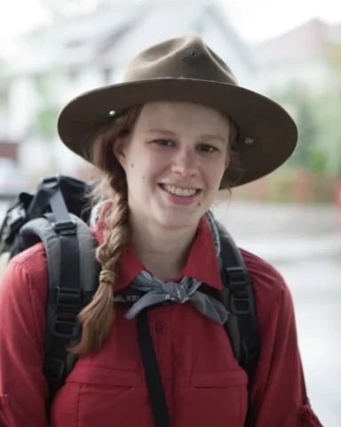 Ranger_IMG_5626 portrait of a woman with light skin and long, light brown hair in a braid. She wears a park ranger hat and a bandana and carries a large backpack.