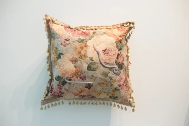 SLRogers_5553_decorative_arts A tan throw pillow hangs on a wall. It has ragged tassels around the edges, a floral fabric in dusty pinks, and appliqued earthworms on it.