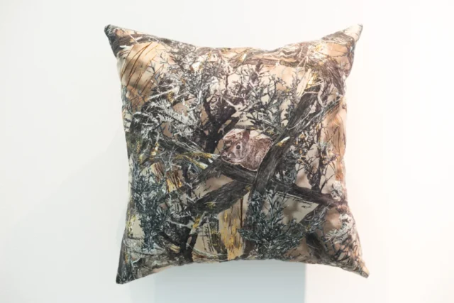 SLRogers_5560_Decorative_Arts A throw pillow hangs on the wall. The fabric is a sage bruch camouflage fabric. Reverse applique techniques shows a rabbit at the center of the pillow.