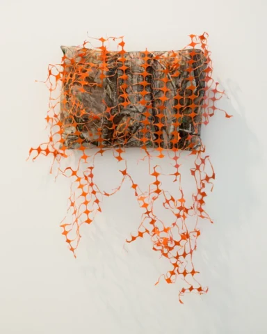 SLRogers_5564_decorative-arts A pillow made with camouflage fabric is covered in torn and tangled orange construction fencing.