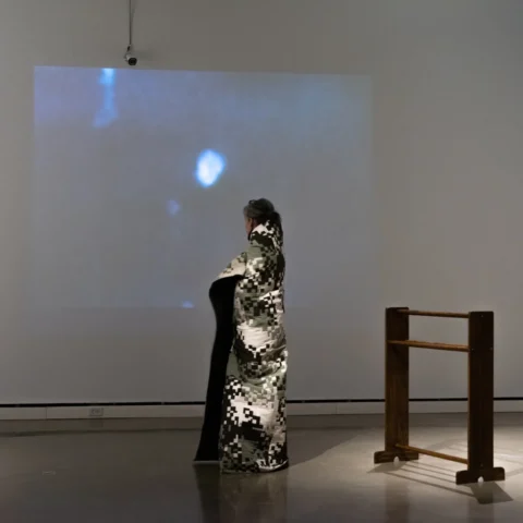 Security_Blanket_DSC00082 A person wraps the artwork "Security Blanket" around their shoulders and turns to look at the projected security camera feed. Only their head is visible on the thermal image. The empty quilt stand is in the corner.