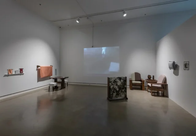 aov9_stephanierogers_dsc0104_45122928754_o A gallery installation view showing a two chairs in a corner opposite a worktable with stools. There is a projected black and white image on the wall and a quilt made in a digital camouflage pattern in the foreground