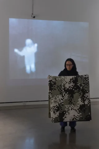 aov9_stephanierogers_dsc0908_45846796731_edit2 A woman holds a camouflage patterned, pieced quilt in front of her. In the background of the photo is a security camera and projected thermal image on the wall, in which the woman appears bright white against a dark gray background. Installation view of "Security Blanket" by Stephanie Lynn Rogers