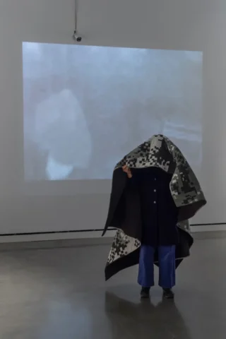 aov9_stephanierogers_dsc0918_45846769711_edit A person drapes a quilt pieced in a digital camouflage pattern over themself, obscuring their image from the thermal security camera in the background and the live video projected onto the wall.
